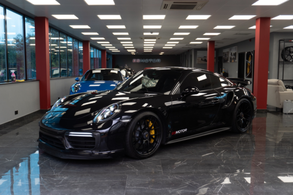 Black Porsche ES900GT in a showroom with a blue car in the background.