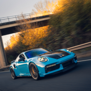 Blue porsche sports car driving on a road with a bridge and autumn trees in the background.