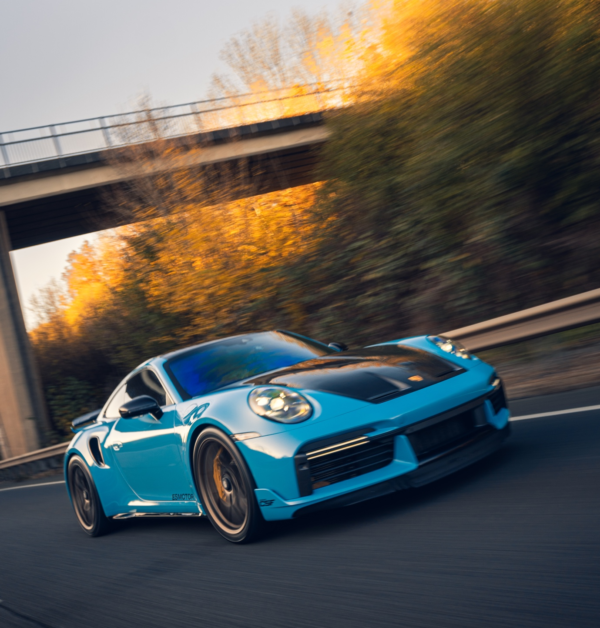 Blue porsche sports car driving on a road with a bridge and autumn trees in the background.