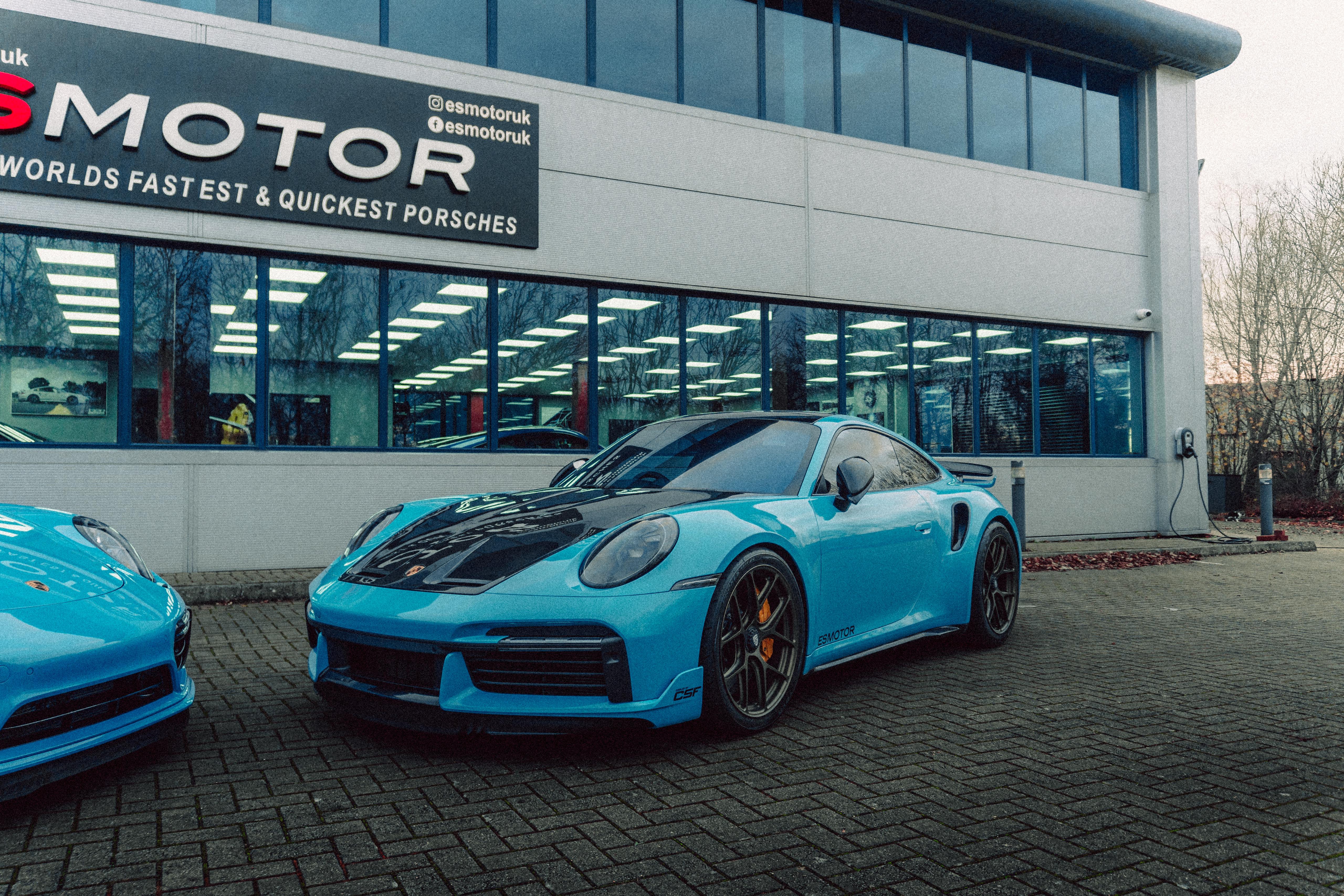 Blue Porsche cars in front of ESMotor building with large windows.