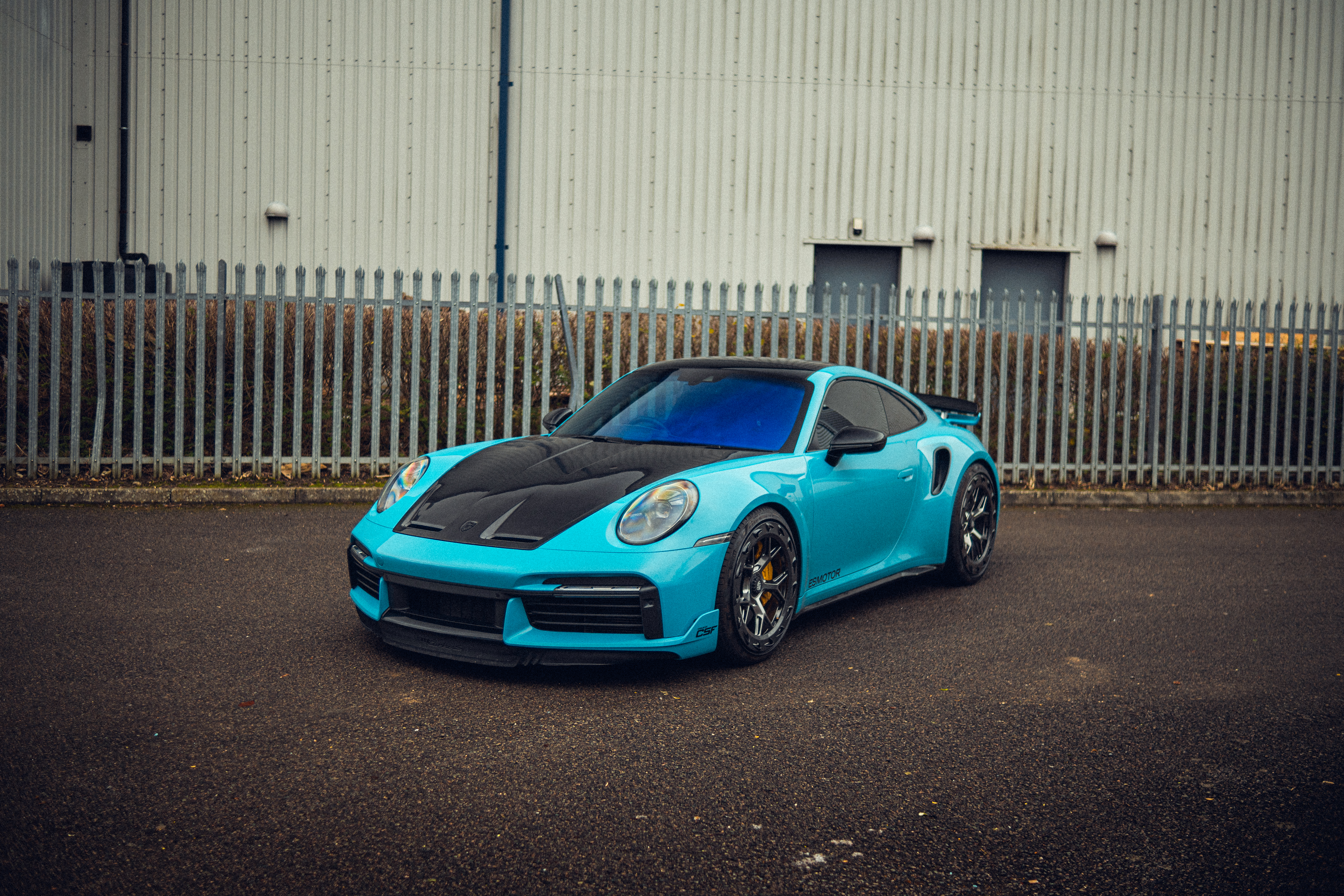 Aqua blue porsche sports car with black hood and custom wheels parked in front of a metal fence and industrial building.